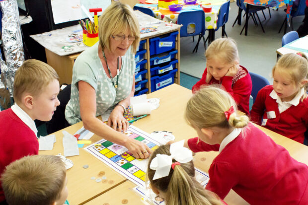 Female teacher sat at a table with primary school pupils explaining a piece of work.