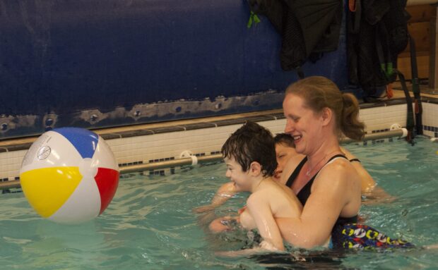 Little boy in the pool with their teacher with the teacher supporting the boy to play with a floating ball.