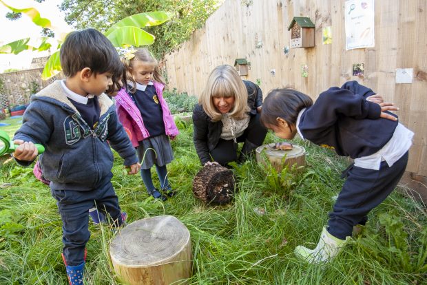 Children watching their teacher intentently studying some bugs outside in the garden under a wooden stump