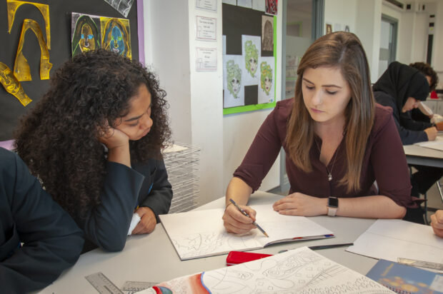 A teacher and a student sitting at the desk where the teacher is explaining something by writing something with her pen in had and a notebook and the student is focused trying to understand