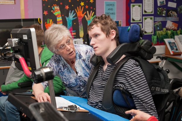 Teenager in a wheelcheer uses a computer with a teacher sat alongside him.