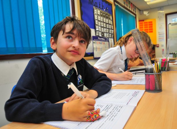 a boy at primary school sits at a desk, writing in his book. Another female classmate sits at a desk next to him. 