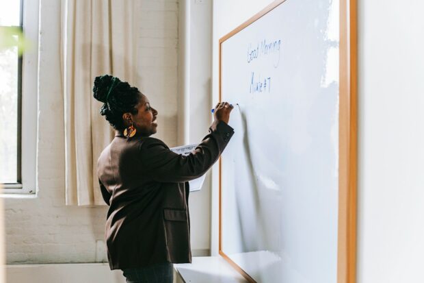 A female Black teacher wearing a suit and writing on a board in a classroom. She is smiling.