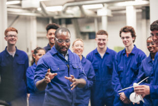 A tutor teaches his class about renewable energy in an engineering workshop. They are all wearing protective eyewear and blue coveralls.