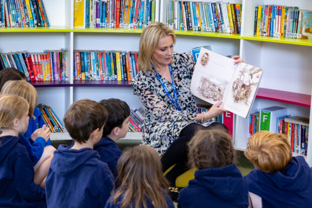 A teacher is reading to a group of young children and showing them the words and pictures in the book. The class is listening attentively. There are shelves of other books in the background.