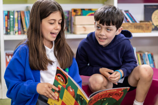 Two children reading a book together in a school library area.